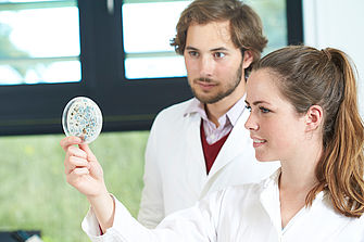 Eine Studentin im Laborkittel hält eine Petri-Schale mit Schimmelsporen hoch. Ein Student schaut ihr über die Schulter. © Hochschule Geisenheim / pps-studios.com