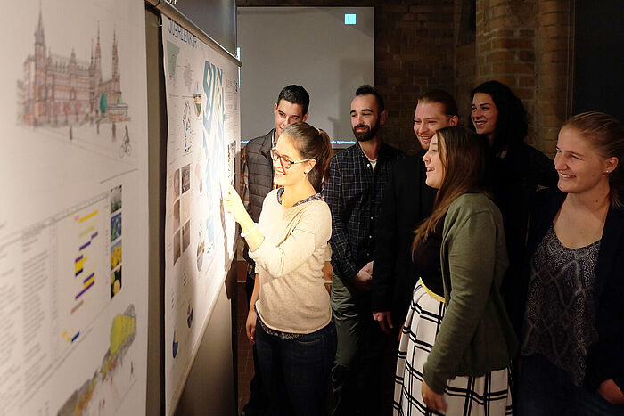 Eine Gruppe Studierender steht vor einer Stellwand mit Plänen und erläutert diese. © Diether von Goddenthow
