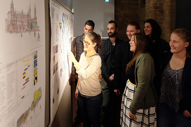 Eine Gruppe Studierender steht vor einer Stellwand mit Plänen und erläutert diese. © Diether von Goddenthow