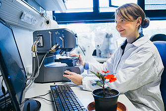 Eine junge Frau im Laborkittel sitzt vor einem Mikroskop, das an einen Bildschirm angeschlossen ist. Neben ihr steht eine rot blühende Pflanze.  © Hessen schafft Wissen / Steffen Boettcher