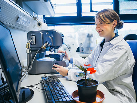 Eine junge Frau im Laborkittel sitzt vor einem Mikroskop, das an einen Bildschirm angeschlossen ist. Neben ihr steht eine rot blühende Pflanze.  © Hessen schafft Wissen / Steffen Boettcher