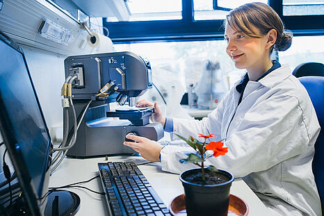 Eine junge Frau im Laborkittel sitzt vor einem Mikroskop, das an einen Bildschirm angeschlossen ist. Neben ihr steht eine rot blühende Pflanze.  © Hessen schafft Wissen / Steffen Boettcher