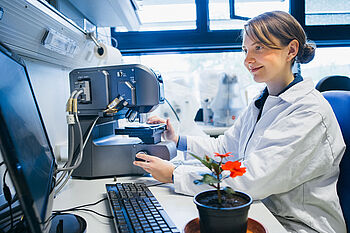 Eine junge Frau im Laborkittel sitzt vor einem Mikroskop, das an einen Bildschirm angeschlossen ist. Neben ihr steht eine rot blühende Pflanze.  © Hessen schafft Wissen / Steffen Boettcher