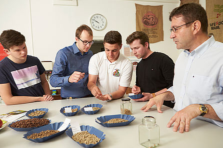 Ein Dozent steht mit einer Gruppe Studenten um einen Tisch. Sie schauen sich unterschiedliche, nicht geröstete Kaffeebohnen an. © Hochschule Geisenheim / pps-studios.com