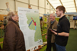 Student und alte Dame und alter Herr stehen vor Plakat