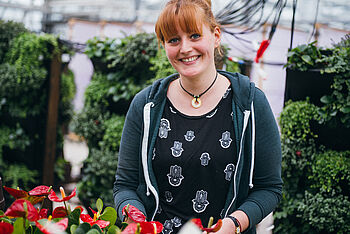 Eine junge Frau steht in einem Gewächshaus, im Hintergrund sind mobile begrünte Wände, im Vordergrund rot blühende Pflanzen. © Hessen schafft Wissen – Steffen Böttcher