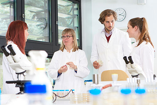 Eine Dozentin steht mit einer Gruppe von Studierenden im Labor. Alle tragen Laborkittel. © Hochschule Geisenheim / pps-studios.com
