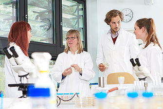 Eine Dozentin steht mit einer Gruppe von Studierenden im Labor. Alle tragen Laborkittel. © Hochschule Geisenheim / pps-studios.com