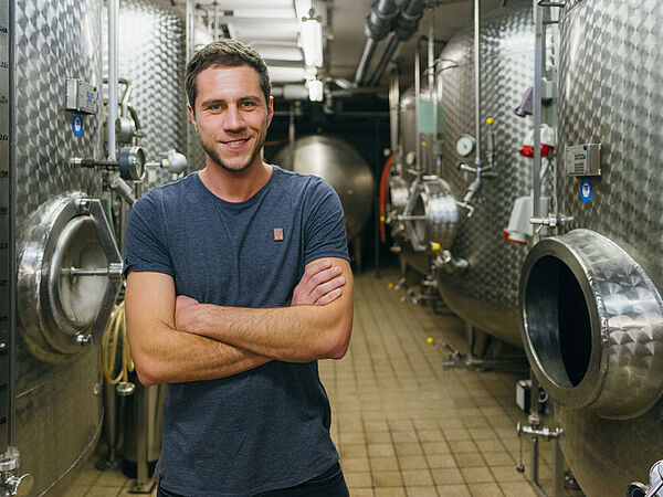 Ein junger Mann im blauen T-Shirt steht vor Edelstahlfässern in einem Weinkeller © Hessen schafft Wissen – Steffen Böttcher 