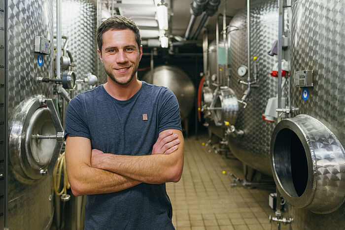 Ein junger Mann im blauen T-Shirt steht vor Edelstahlfässern in einem Weinkeller © Hessen schafft Wissen – Steffen Böttcher 