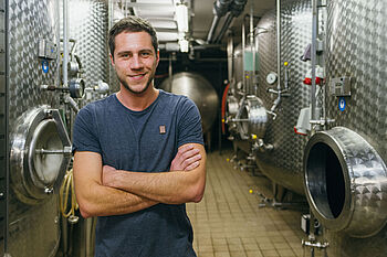 Ein junger Mann im blauen T-Shirt steht vor Edelstahlfässern in einem Weinkeller © Hessen schafft Wissen – Steffen Böttcher 