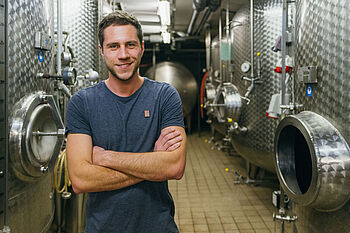 Ein junger Mann im blauen T-Shirt steht vor Edelstahlfässern in einem Weinkeller © Hessen schafft Wissen – Steffen Böttcher 