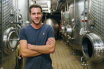 Ein junger Mann im blauen T-Shirt steht vor Edelstahlfässern in einem Weinkeller © Hessen schafft Wissen – Steffen Böttcher 