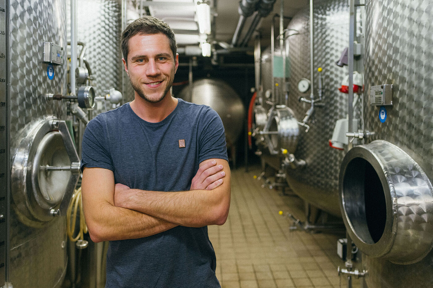 Ein junger Mann im blauen T-Shirt steht vor Edelstahlfässern in einem Weinkeller © Hessen schafft Wissen – Steffen Böttcher