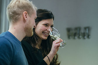 Eine junge Frau mit dunklen Haaren riecht an einem Glas mit Wein, ein blonder junger Mann steht daneben © Hessen schafft Wissen – Steffen Böttcher 
