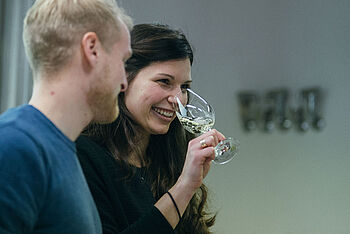 Eine junge Frau mit dunklen Haaren riecht an einem Glas mit Wein, ein blonder junger Mann steht daneben © Hessen schafft Wissen – Steffen Böttcher 