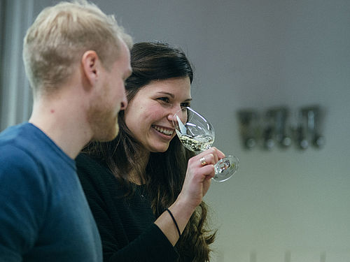 Eine junge Frau mit dunklen Haaren riecht an einem Glas mit Wein, ein blonder junger Mann steht daneben © Hessen schafft Wissen – Steffen Böttcher 