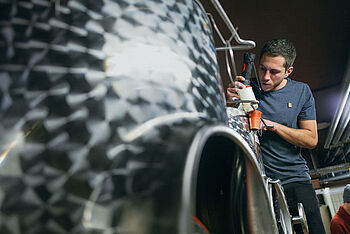 Ein junger Mann steht an einem Stahltank im Weinkeller. © Hessen schafft Wissen – Steffen Böttcher