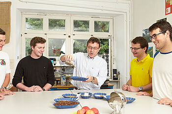 Ein Dozent schüttet Kaffee-Bohnen aus einem Glas in eine Schale. Um ihn herum stehen Studierende und schauen zu. © Hochschule Geisenheim / pps-studios.com