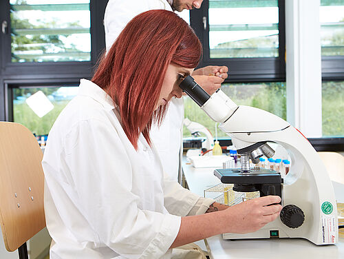 Eine rothaarige Studierende im Laborkittel schaut durch ein Mikroskop © Hochschule Geisenheim / ppsstudios.com