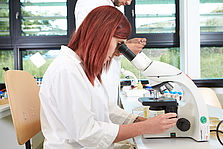 Eine rothaarige Studierende im Laborkittel schaut durch ein Mikroskop © Hochschule Geisenheim / ppsstudios.com