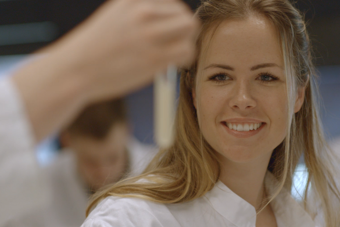 Eine junge Frau im Laborkittel betrachtet den Inhalt eines Reagenzglases. © Hochschschule Geisenheim