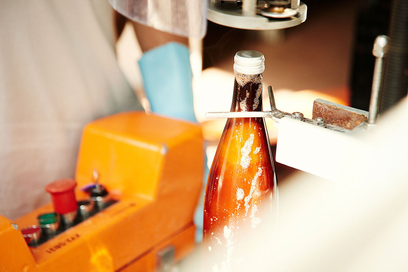 Eien Person mit Handschuhen steht vor einem Gerät, das Flaschen nach der Abfüllung verschließt. © Hessen schafft Wissen / Daniel Chassein