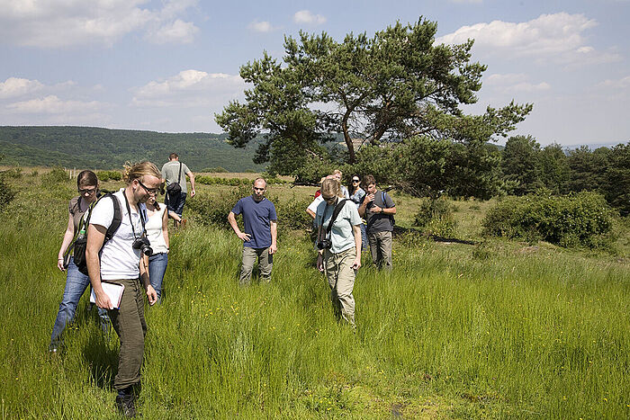 Studierende mit Rucksäcken stehen auf einer Wiese mit hohem Gras und betrachten ihre Umgebung. © Hochschule Geisenheim