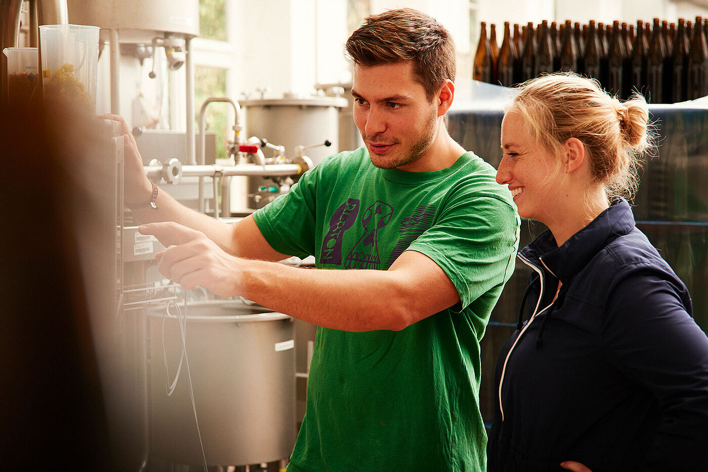 Ein junger Mann erklärt einer jungen Frau eine technische Anlage. Im Hintergrund stehen leere Flaschen. © Hessen schafft Wissen / Daniel Chassein