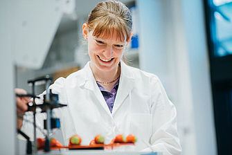 Eine Frau im Laborkittel steht vor einer Apparatur auf der Erdbeeren auf einer schwarzen Platte liegen © Hessen schafft Wissen – Steffen Böttcher 