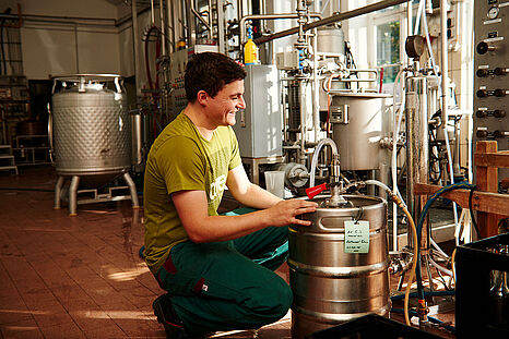 Ein junger Mann hockt vor einem Keg an einer Abfüllanlage. © Hessen schafft Wissen / Daniel Chassein