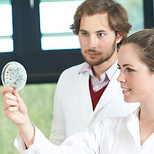 Eine Studentin im Laborkittel hält eine Petri-Schale mit Schimmelsporen hoch. Ein Student schaut ihr über die Schulter. © Hochschule Geisenheim / pps-studios.com