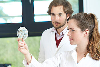 Eine Studentin im Laborkittel hält eine Petri-Schale mit Schimmelsporen hoch. Ein Student schaut ihr über die Schulter. © Hochschule Geisenheim / pps-studios.com